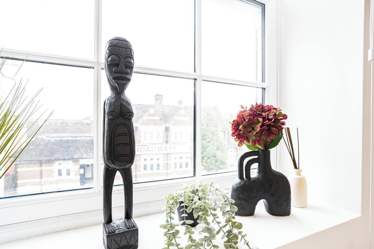 A room with a large window, a black sculpture, a vase with red flowers, and a small plant on a windowsill.