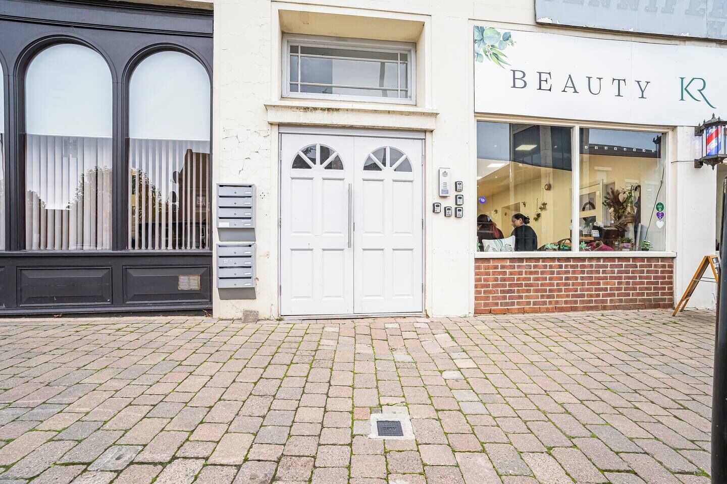 A building with a white facade, a brick entrance, and a storefront with a sign that reads 'BEAUTY R'.