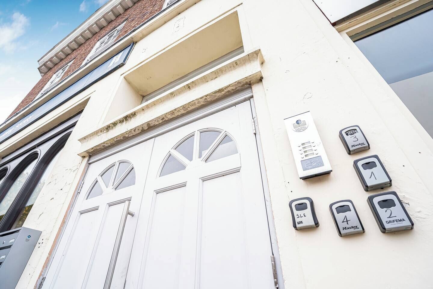 Fachada de edificio con exterior blanco, un juego de puertas dobles con ventanales arqueados y una fila de botones de llamada numerados a la derecha.