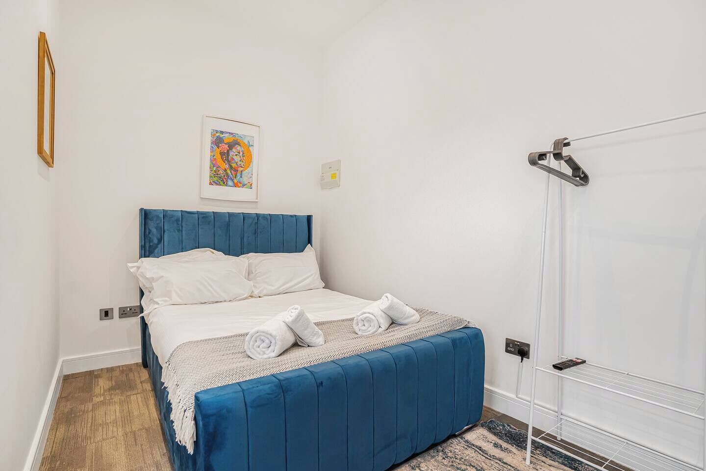 Un dormitorio con cabecera tapizada en azul, una cama con ropa blanca, una toalla enrollada sobre la cama y un estante en la pared con un libro.