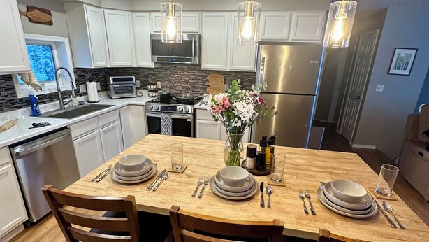 Une cuisine moderne avec des meubles blancs, des appareils en acier inoxydable et une table à manger en bois pour quatre personnes.