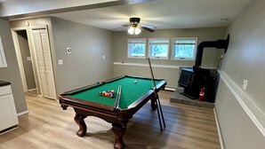 Une table de billard dans une pièce au plancher en bois, avec un ventilateur de plafond et une fenêtre équipée de stores.