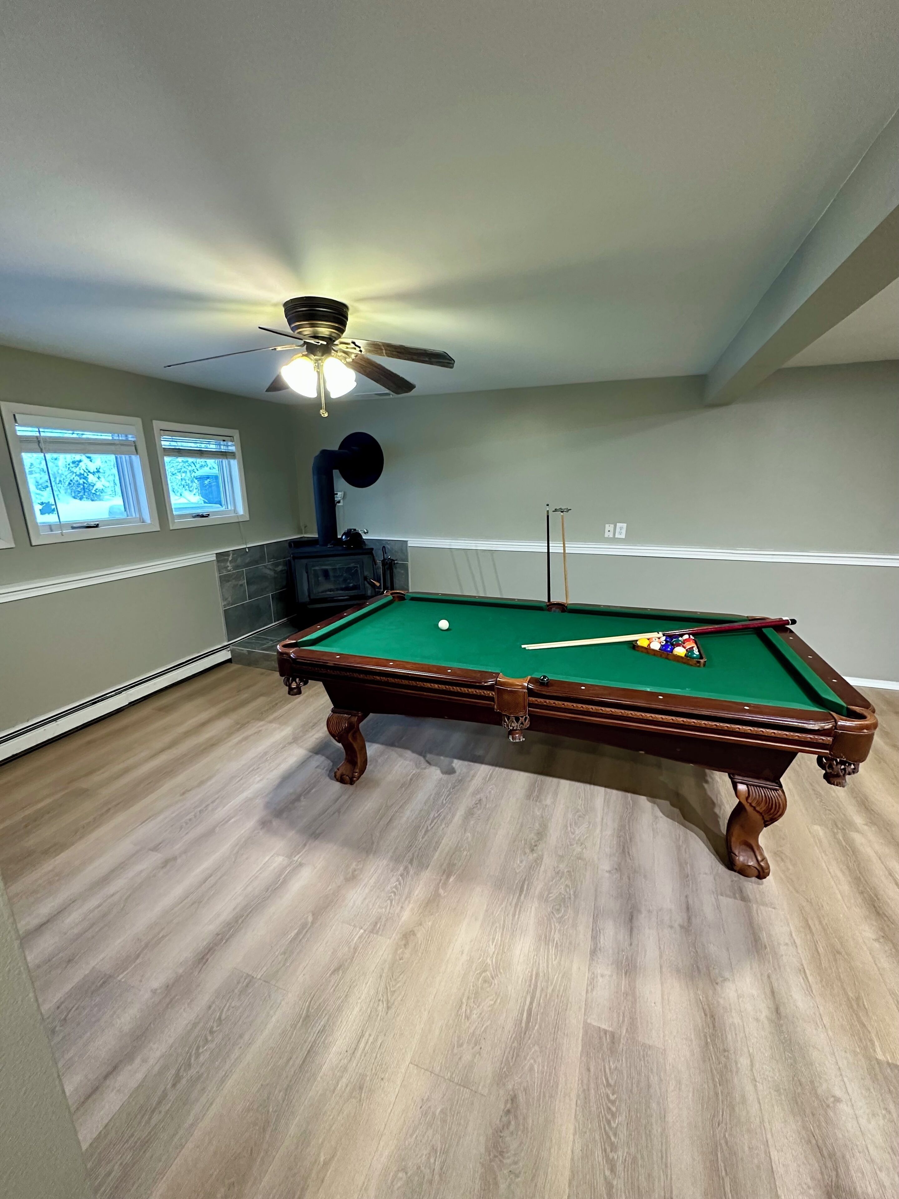 Una mesa de billar en una habitación con ventilador de techo, chimenea y ventanas con persianas.
