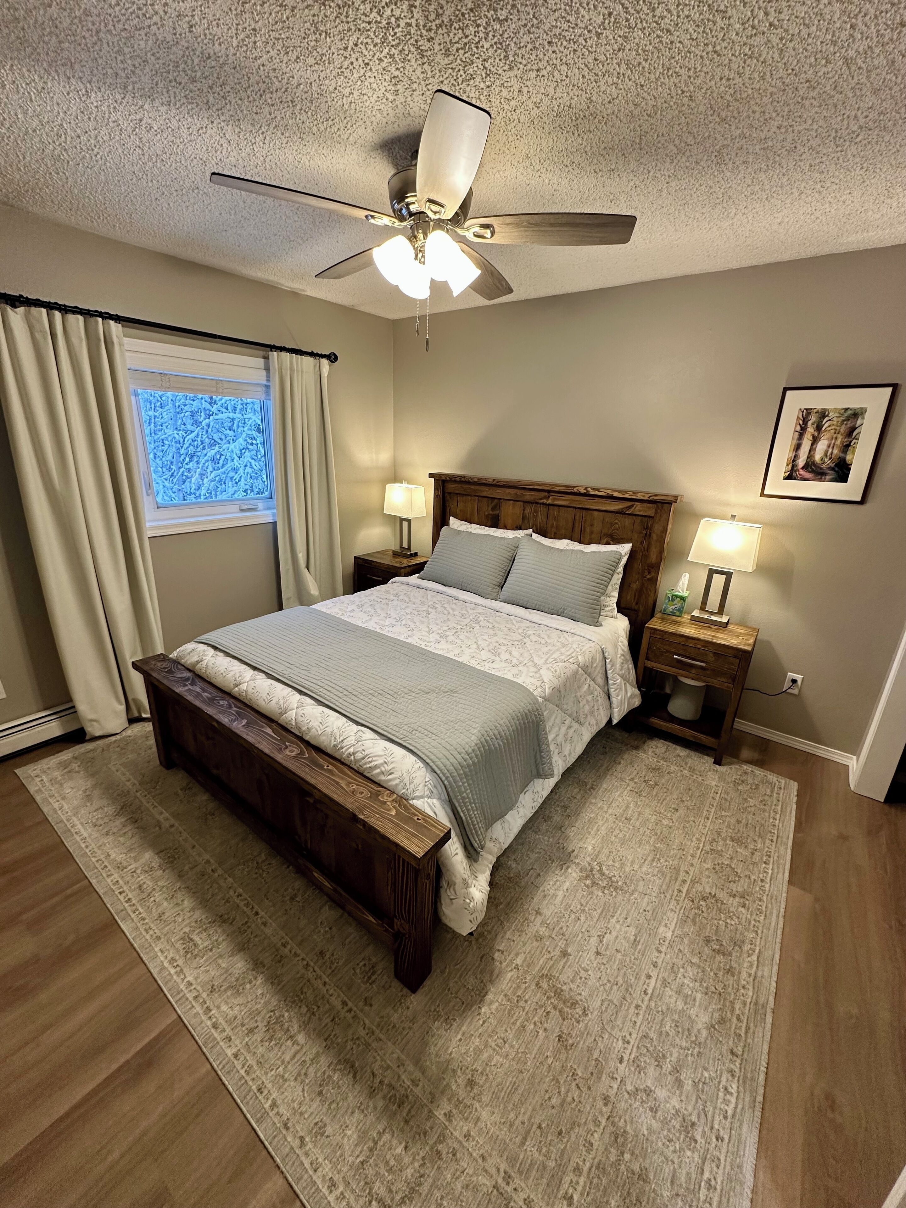 A bedroom with a bed, two bedside tables with lamps, a ceiling fan, a window with curtains, and a framed picture on the wall.