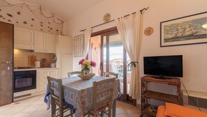 Una cocina con armarios blancos, una mesa de comedor con sillas, un televisor sobre un mueble de madera y un balcón con cortinas.