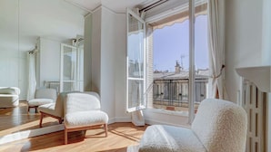 Ein geräumiges Zimmer mit Holzböden, einem großen Fenster mit Blick auf Gebäude und zwei weißen Sesseln.