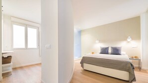 Un dormitorio con cama, mesitas de noche, una ventana y una lámpara fijada a la pared.