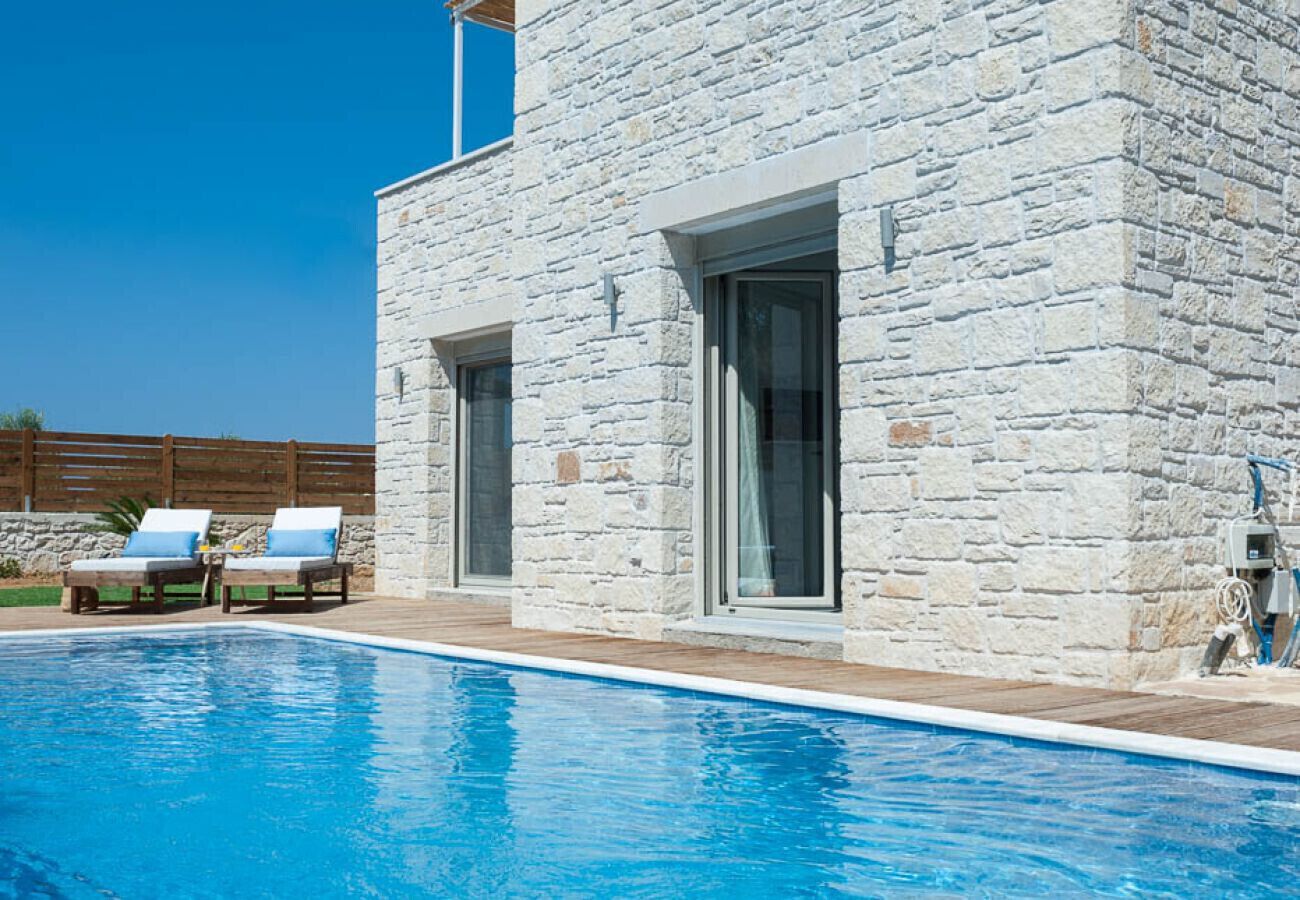 A modern stone building with a swimming pool, two lounge chairs, and a wooden fence in the background.