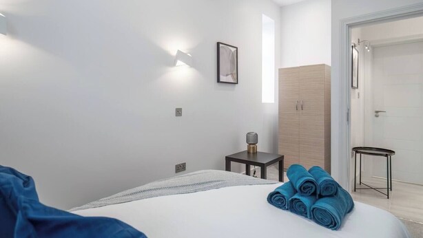 Moderni hotellihuone, jossa on sänky, pieni pöytä, seinään kiinnitetty valaisin ja ovi, joka johtaa kylpyhuoneeseen.
