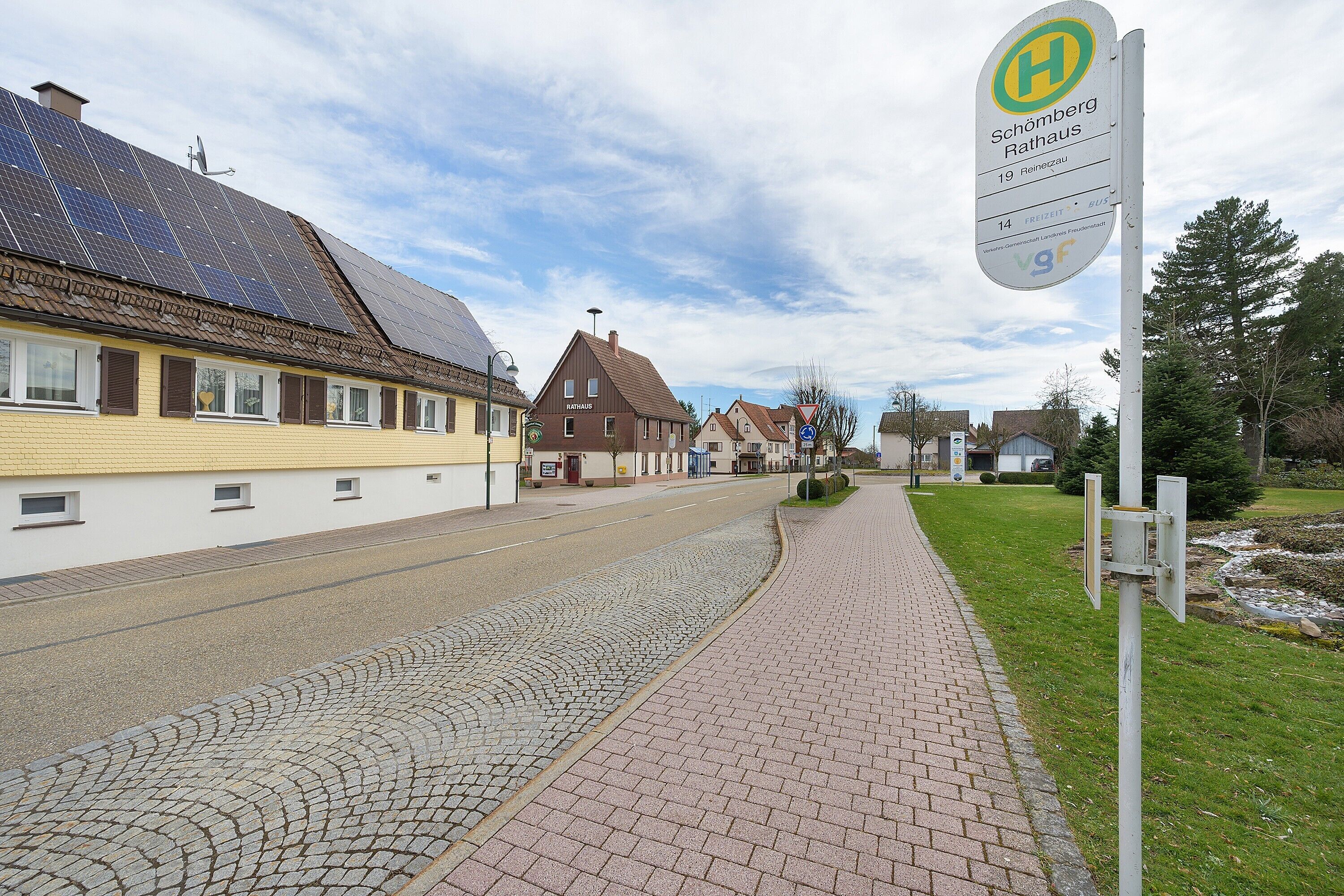 Une rue bordée de bâtiments dont les toits sont recouverts de panneaux solaires, une enseigne indiquant « Schömburg Rathaus 19 Hennegau », et un chemin pavé.
