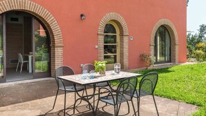 A patio with a table and chairs, a small garden, and a building with arched windows and a red facade. - Apartment Azzurro- Cignella Resort Tuscany (Trequanda)