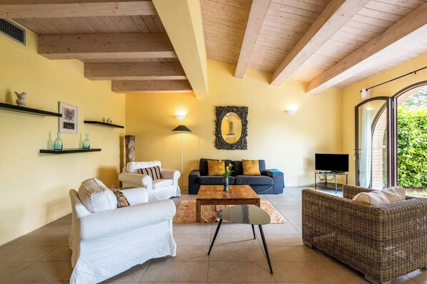A living room with a sofa, a coffee table, and a wicker sofa. There is a TV on a stand and a window with a view of greenery. - Villa Limone - 3 Bedroom Villa in Tuscany (Trequanda)
