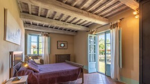 Een slaapkamer met een houten plafond, een bed met een rode deken, een nachtkastje, een dressoir en een raam met gordijnen.