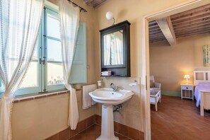 A bathroom with a white pedestal sink, a mirror above it, and a window with curtains. There is a towel rack and a small table with a lamp. - Apartment Olivo - 3 Bedroom Apartment in Tuscany (Trequanda)