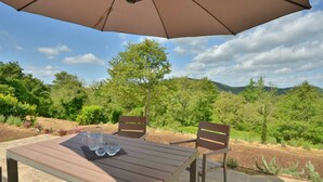 Une terrasse extérieure avec une table et des chaises, un parasol, et une vue sur un jardin aménagé avec des arbres et des collines.