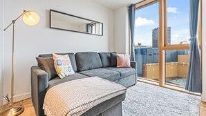 A modern living room with a grey sofa, a floor lamp, and a large window with a view of buildings. - Apt-sleeps 2/3-bullring & Grand Central Close (Birmingham)