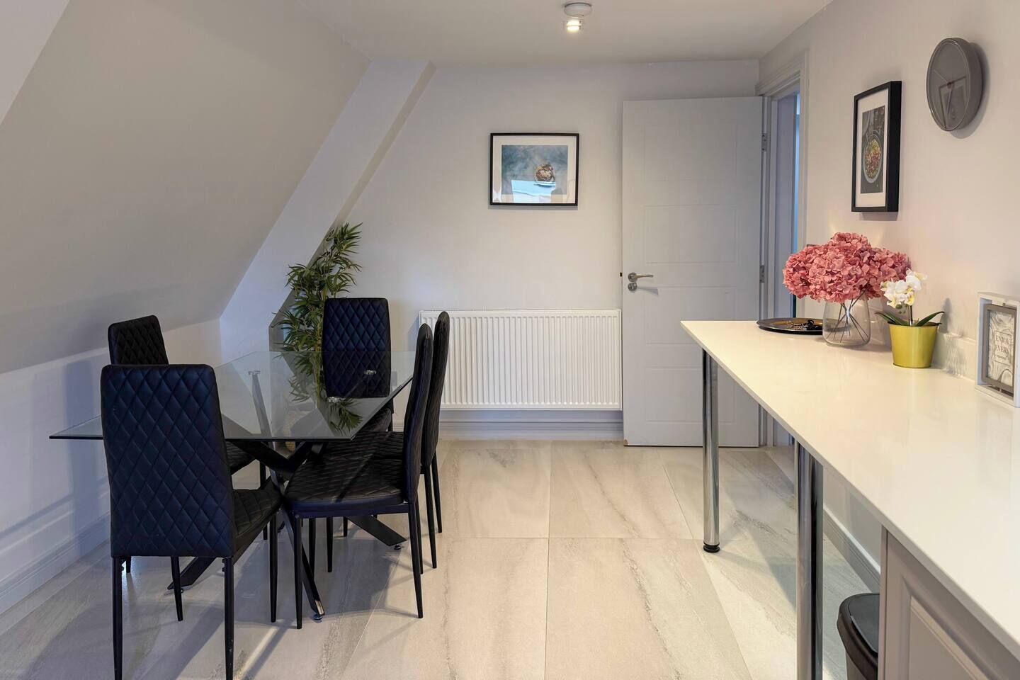 Een moderne keuken met een wit aanrecht, zwarte stoelen en een potplant.
