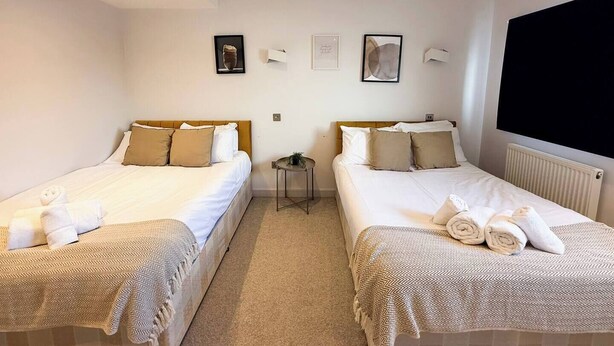 A hotel room with two beds, each with a patterned bedspread and pillows, a small side table, and a wall-mounted TV. - Contemporary 2 Bed Apt in Didcot (Sleeps 7) (Didcot)