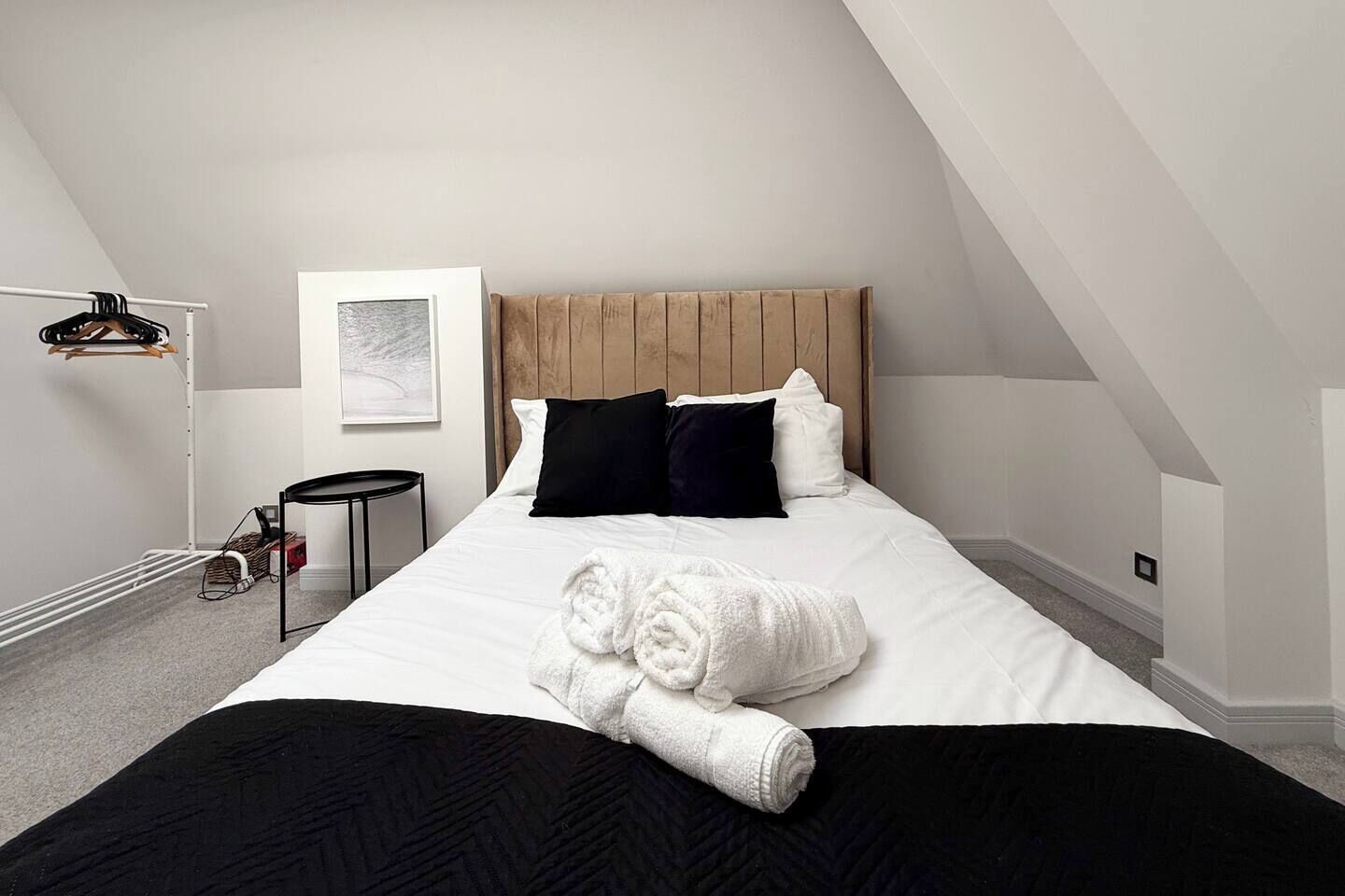 A bedroom with a bed, a wooden headboard, a black and white pillow set, a rolled white towel, a small round table, and a hanging rack.