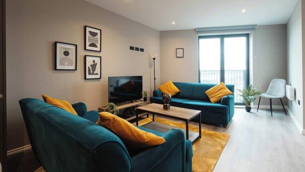 Een moderne woonkamer met blauwgroene banken, een salontafel en een televisie. Er hangen afbeeldingen in lijsten aan de muur en een groot raam met zonwering.