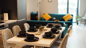 Een eetgedeelte met een lange tafel gedekt voor een maaltijd, een bank met gele kussens, en een uitzicht op de stad door grote ramen.