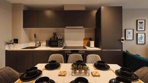 Een moderne eetruimte met een witte tafel voor vier personen, donkergrijze stoelen en een keuken op de achtergrond.