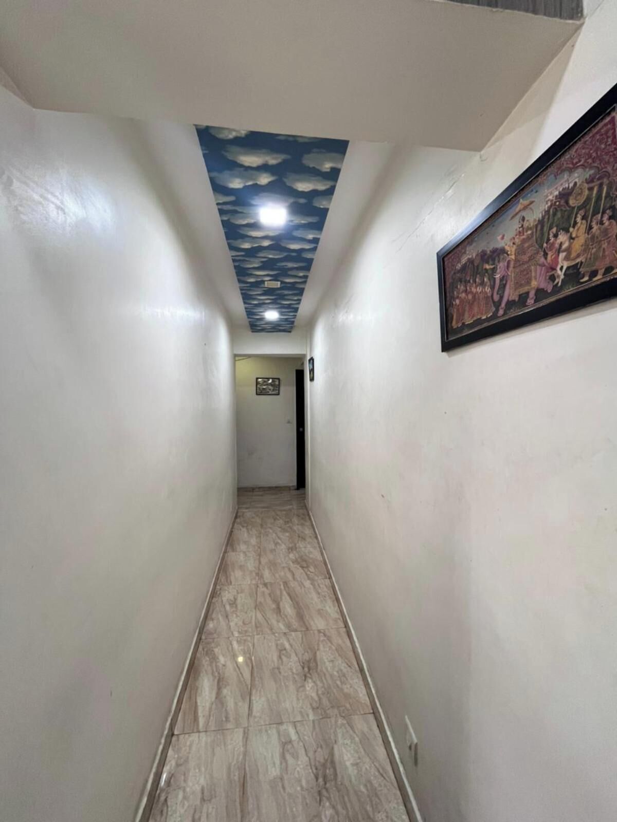 Un couloir dont le plafond est peint pour ressembler à des nuages, avec un sol carrelé et un tableau encadré accroché au mur.
