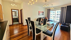Uma sala de estar moderna com uma mesa de jantar de vidro, cadeiras preto e branco, um sofá preto e uma televisão. Há uma janela com cortinas e vista para edifícios.