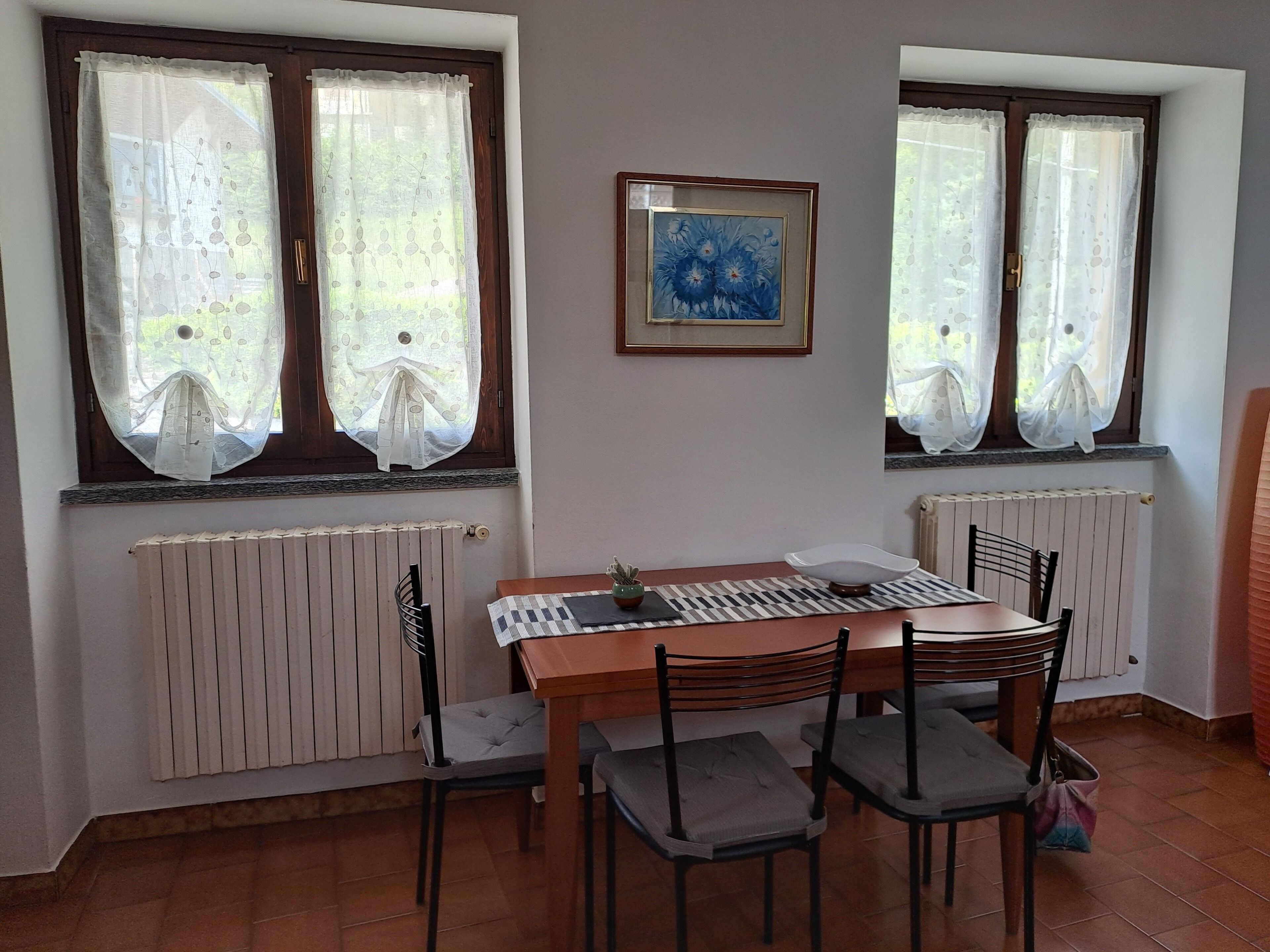 Un comedor con una mesa y sillas, dos ventanas con cortinas, un radiador y un cuadro en la pared.