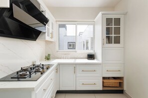 Une cuisine avec des meubles blancs, une cuisinière noire et une fenêtre avec des stores.