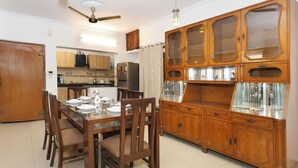 A dining area with a wooden table and chairs, a kitchen in the background, and a large wooden cabinet with glass doors. - Amber Crest by Redolive10min ~airportdelhi2bhk (New Delhi)