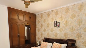 A bedroom with a wooden bed, a ceiling fan, a wardrobe, and patterned wallpaper. - Amber Crest by Redolive10min ~airportdelhi2bhk (New Delhi)