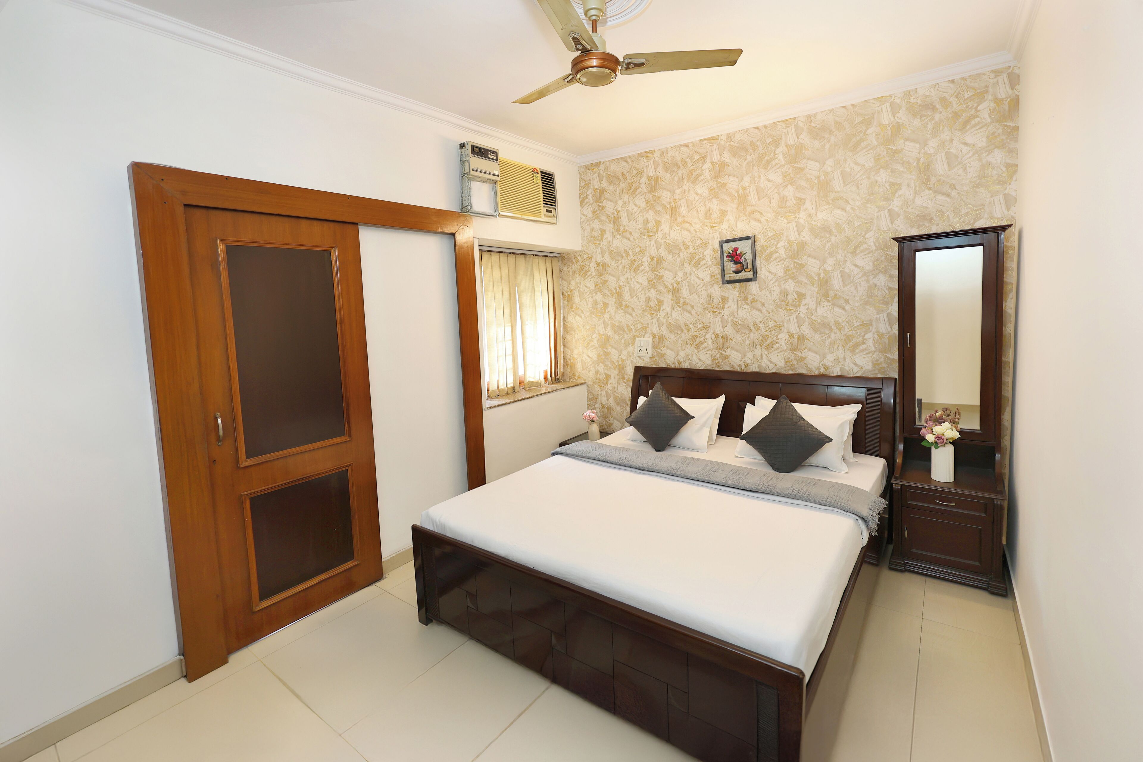 A bedroom with a bed, a wooden wardrobe, a nightstand, a mirror, a ceiling fan, and an air conditioning unit.