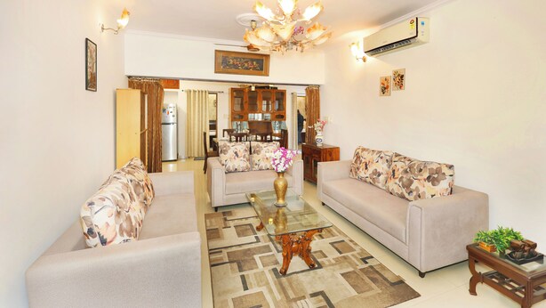 A living room with two sofas, a glass coffee table, a chandelier, and an air conditioning unit. - Amber Crest by Redolive10min ~airportdelhi2bhk (New Delhi)