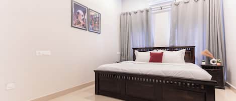 Un dormitorio con una cama de madera oscura, ropa de cama blanca, una mesita de noche con lámpara y una ventana con cortinas.