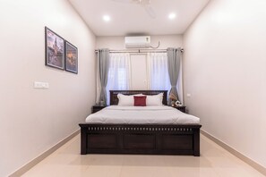 Une chambre avec un grand lit, un ventilateur de plafond, un appareil de climatisation et deux tableaux encadrés au mur.