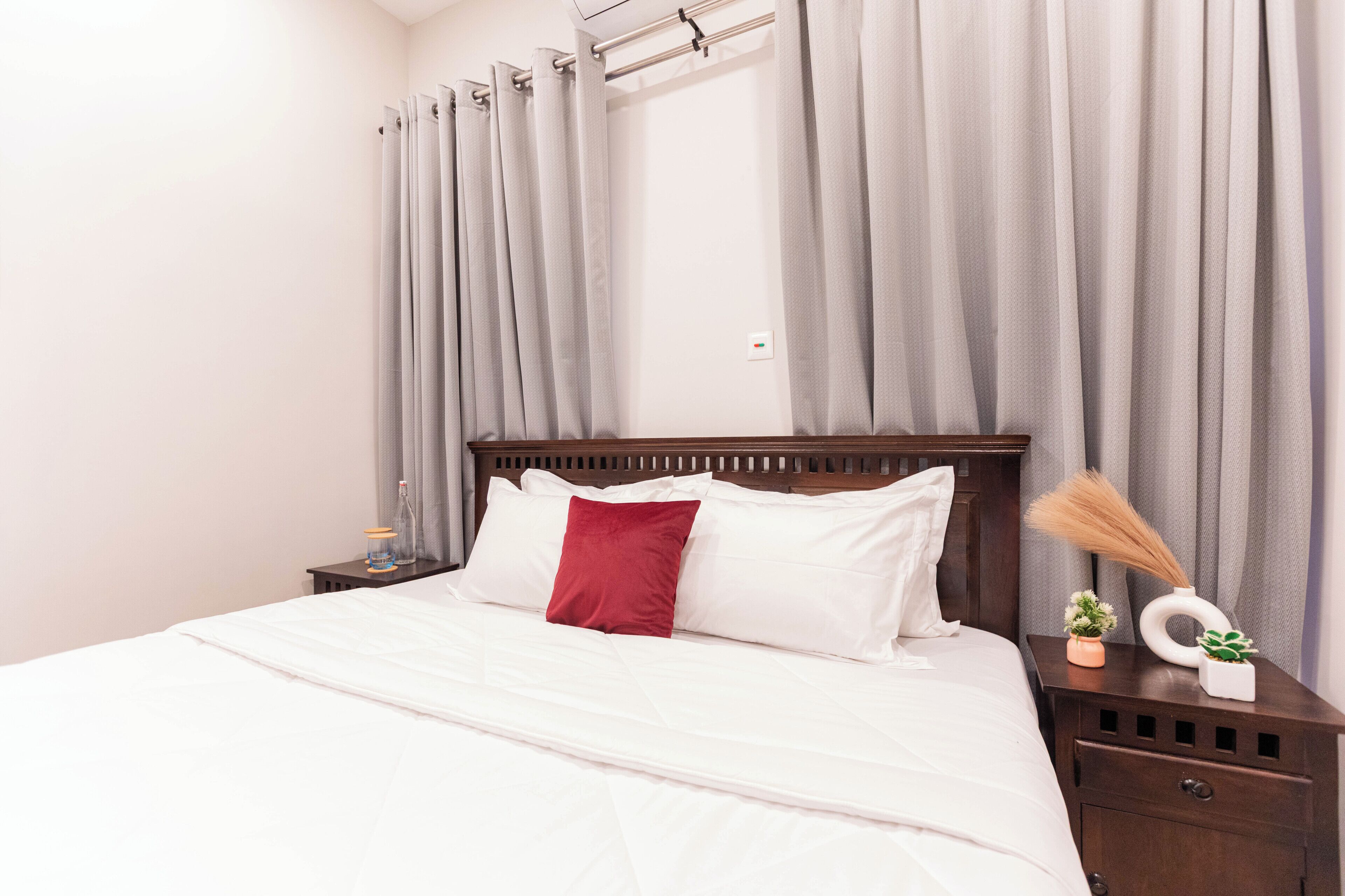 A neatly made bed with white and red bedding, a wooden headboard, and a nightstand with a small plant and a decorative item.