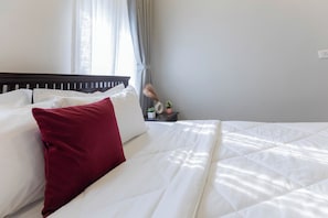 A neatly made bed with white and red pillows, a dark headboard, and a bedside table with a small plant and a clock.