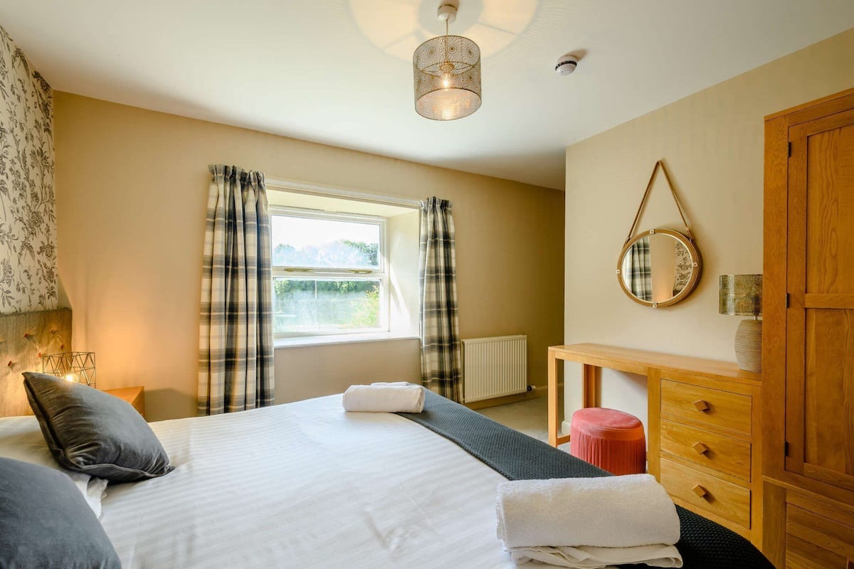 A well-lit bedroom with a bed, a bedside table, a mirror, and a window with curtains.