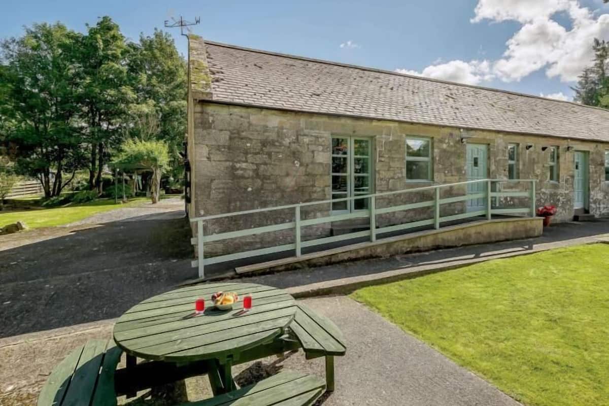 A stone building with a sloped roof, a covered outdoor seating area, and a picnic table with food and drinks.
