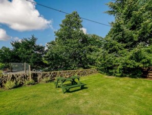 A green picnic table in a grassy backyard with a stone wall and trees in the background.