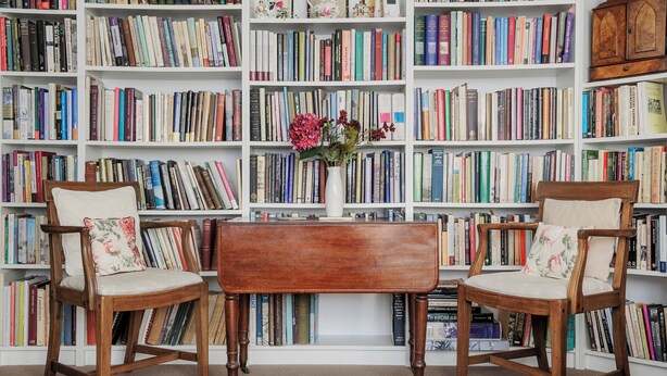 Een kamer met twee houten stoelen, een klein houten tafeltje en een boekenplank vol boeken.