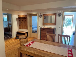 Un comedor con una mesa y sillas de madera, una cocina visible a través de una puerta abierta, y un espejo sobre un armario de madera.