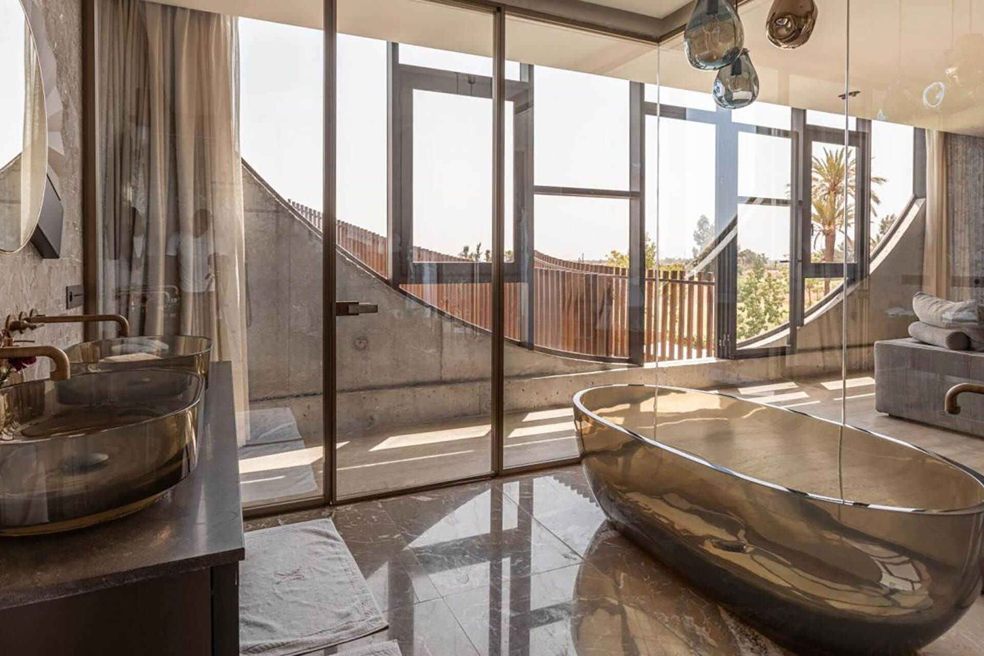 A modern bathroom with a large glass-enclosed bathtub, a vanity with a gold faucet, and a view of a wooden deck outside.
