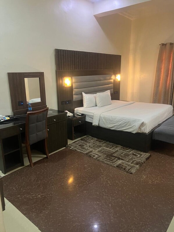A hotel room with a bed, a desk, a chair, a mirror, and a nightstand. - Nippon Grand Hotel and Apartment (Abuja)