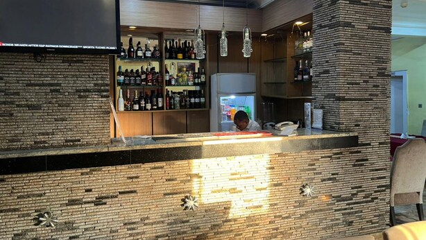 Een bar met een bakstenen muur, drankflessen op planken, hangende lampen en een stenen pilaar.