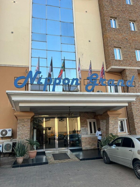 La façade extérieure du Nippon Grand Hotel, avec sa grande surface vitrée, un bâtiment en pierre sur la droite et de nombreux drapeaux déployés à l’extérieur.