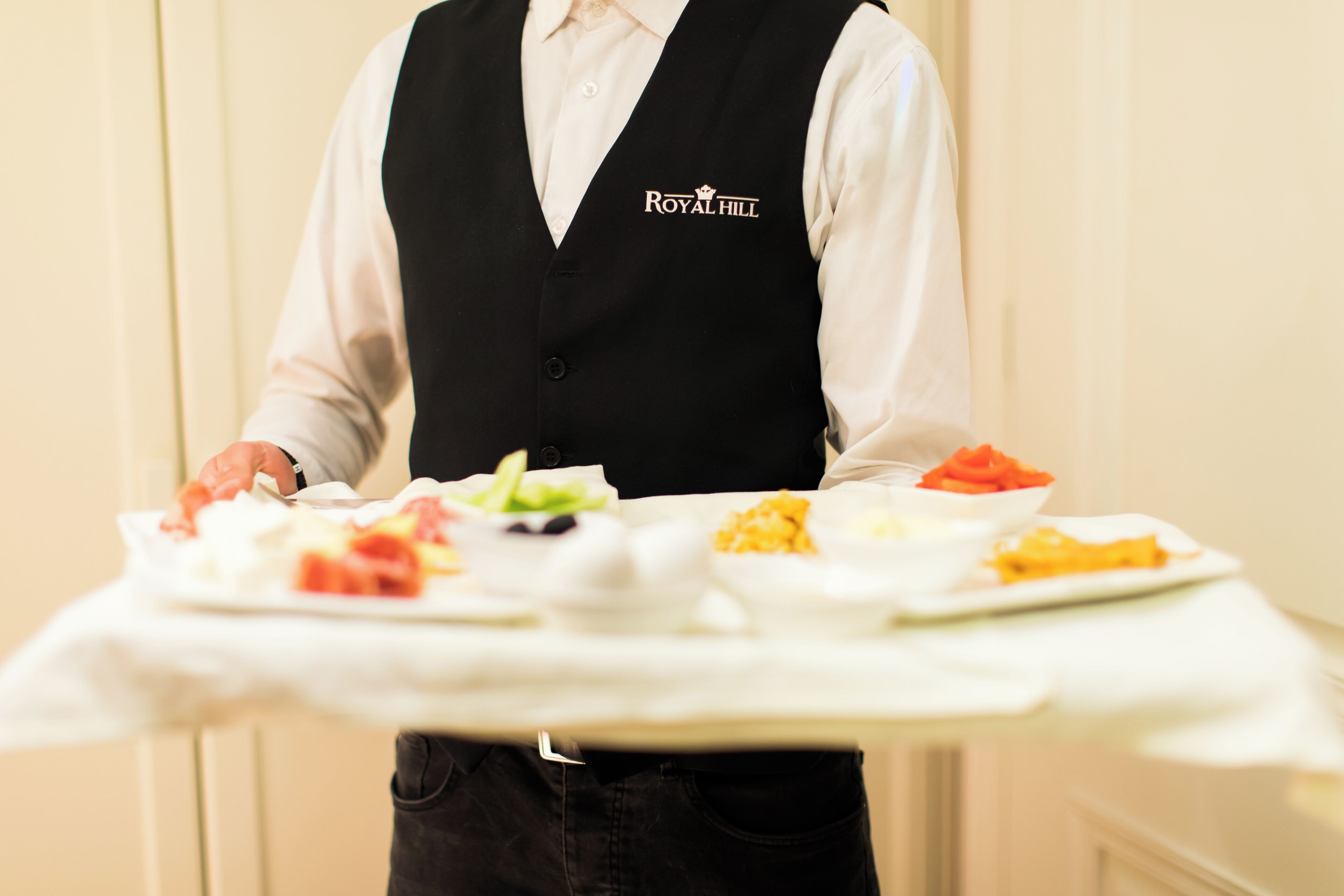 A person in a black vest with "ROYAL HILL" logo carrying a tray with various dishes.