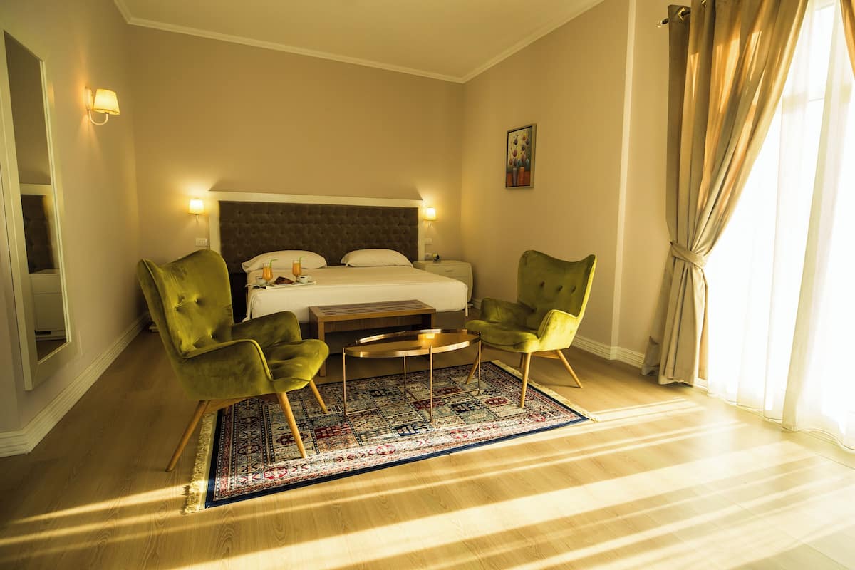 Une chambre d’hôtel avec un lit, deux fauteuils verts, une petite table, un tapis à motifs et une grande fenêtre avec des rideaux.