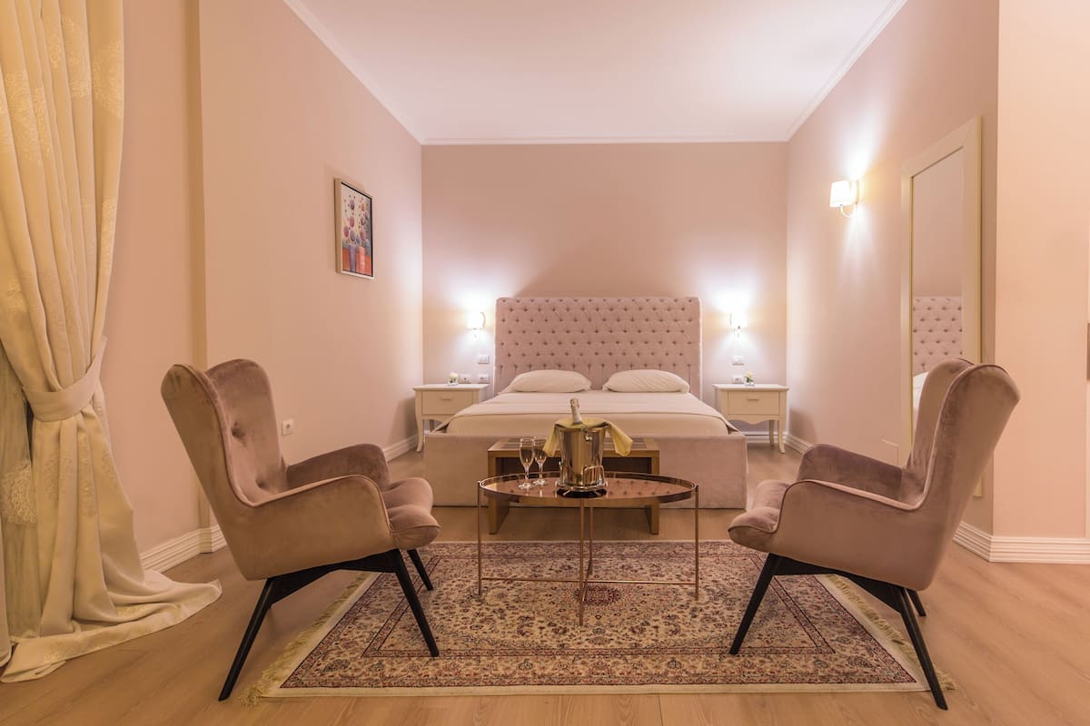 Une chambre d’hôtel avec un grand lit, deux fauteuils, une petite table avec une bouteille et des verres, un tapis et un tableau au mur.